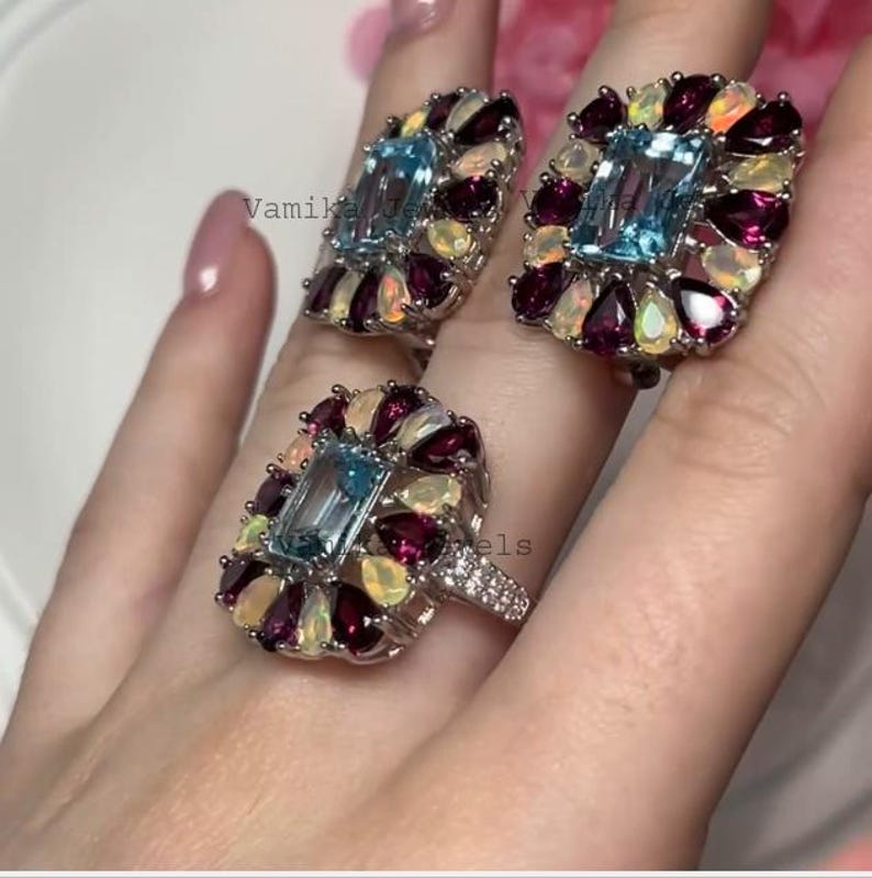 May include: A set of three gemstone rings and earrings. Each piece features a rectangular, light blue gemstone surrounded by smaller, colorful gems in a floral arrangement. The rings and earrings are set in silver.
