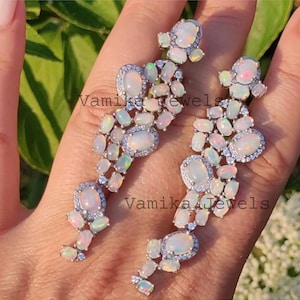 May include: A pair of long, dangling earrings featuring oval and round opals, accented with small, clear gemstones. The earrings have a silver-colored setting and the text "Vamika Jewels".