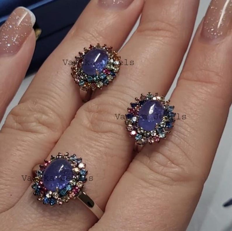 May include: Three silver rings with oval, blue gemstones surrounded by a halo of smaller, colorful stones. The rings are displayed on a hand, showcasing the intricate details of the jewelry. The rings are from Vanka Jewels.