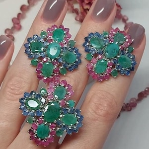 May include: Emerald and gemstone jewellery set. The set includes a ring and two earrings. The jewellery features large, oval-cut emeralds surrounded by blue and pink gemstones. The jewellery is set in a silver metal.