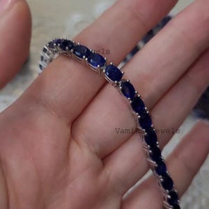 May include: A silver bracelet featuring a series of oval, deep blue gemstones. The gemstones are set closely together, creating a continuous line of color. The bracelet is shown against a soft, neutral background.