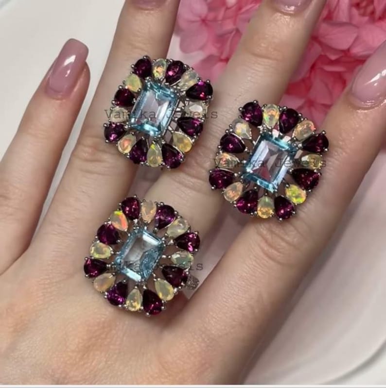 May include: Three rings with a square, light blue gemstone center, surrounded by a halo of smaller, teardrop-shaped gemstones in shades of purple and yellow. The rings are set on a silver band.