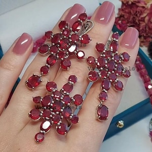 Op de afbeelding: Een set robijnrode oorbellen en een ring, met een bloemmotief. De sieraden zijn gezet met talrijke ovale en ronde edelstenen. De oorbellen en ring worden getoond op een hand met roze nagellak.