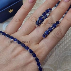 May include: A jewelry set featuring a bracelet, ring, and earrings, all adorned with oval-cut, deep blue gemstones. The pieces are set in a silver-toned metal. The bracelet and earrings are visible, with the ring resting on a hand. The brand name "Vamika Jewels" is visible.
