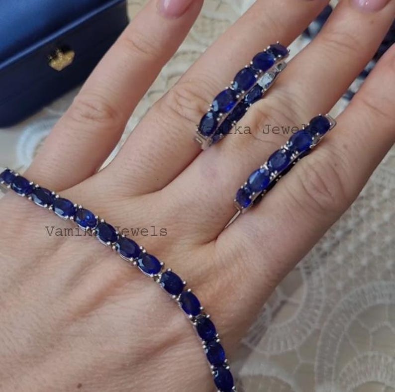 May include: A jewelry set featuring a bracelet, ring, and earrings, all adorned with oval-cut, deep blue gemstones. The pieces are set in a silver-toned metal. The bracelet and earrings are visible, with the ring resting on a hand. The brand name "Vamika Jewels" is visible.