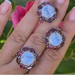 May include: Three silver rings with a floral design. Each ring features a large, round, light-coloured stone surrounded by smaller, oval, dark red stones. The rings have the text "Vamika Jewels" on them.