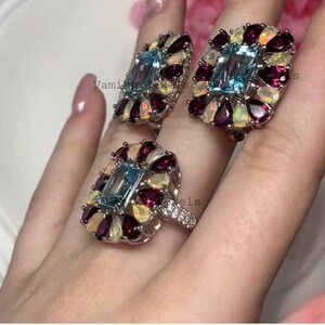 May include: Three silver rings with a rectangular light blue gemstone center, surrounded by smaller, colorful gemstones. The rings are set with a cluster of red, white, and yellow stones. The rings are on a person's finger.