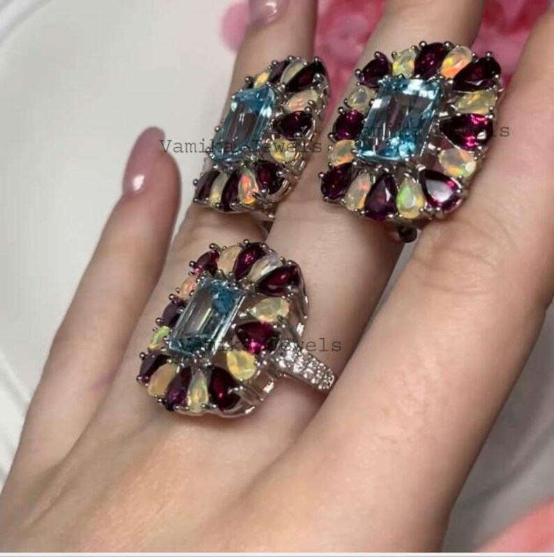 May include: Three silver rings with a rectangular light blue gemstone center, surrounded by smaller, colorful gemstones. The rings are set with a cluster of red, white, and yellow stones. The rings are on a person's finger.