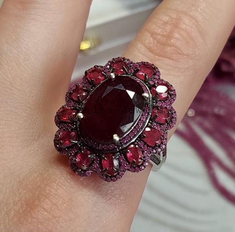 May include: An elaborate ring with a central oval ruby, encircled by smaller rubies. The ring has a silver-toned band and a floral design. The rubies are a deep red, creating a striking contrast. The ring's design is intricate and eye-catching.