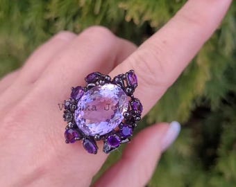 May include: An elaborate ring with a large, oval, pale purple gemstone at its centre, encircled by smaller, dark purple stones. The ring's setting is a dark metal. The ring is on a finger, with a blurred green backdrop.