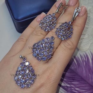 May include: A jewellery set comprising a ring, pendant, and earrings, each piece embellished with clusters of small, oval-shaped, light purple gemstones. The earrings and pendant have a teardrop design. The ring has a similar design. The jewellery is set against a neutral background.