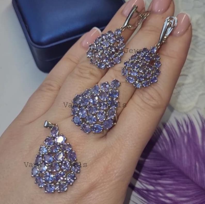 May include: A jewellery set comprising a ring, pendant, and earrings, each piece embellished with clusters of small, oval-shaped, light purple gemstones. The earrings and pendant have a teardrop design. The ring has a similar design. The jewellery is set against a neutral background.
