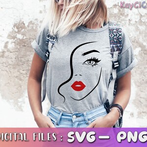May include: A gray t-shirt with a black and white line drawing of a woman's face with red lips. The woman's eye is visible and has a black eyelash. The t-shirt is being worn by a person with blonde hair.