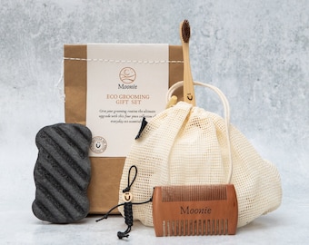 Puede incluir: Una caja de regalo de papel marrón con el texto "Moonie ECO GROOMING GIFT SET" y una pequeña ilustración de una luna. La caja contiene un jabón negro de carbón vegetal, un cepillo de dientes de madera, una pequeña bolsa de malla blanca y un peine de madera con el texto "Moonie" grabado en él.