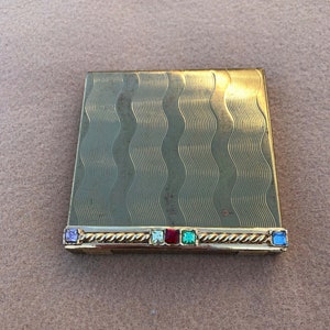 Puede incluir: Un estuche compacto dorado vintage con un patrón ondulado en la superficie. El estuche es rectangular con un borde decorativo con piedras preciosas de colores. Las piedras preciosas son moradas, verdes, rojas y azules.