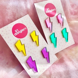 May include: Two sets of earrings on display. Each set features three lightning bolt-shaped earrings in vibrant colors: yellow, pink, purple, and green. The earrings are displayed on white cards with the text "li'l danglers" in a pink circle.