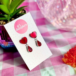 May include: Red heart and teardrop earrings on a white card. The earrings are suspended from silver-coloured metal. The card has the text "li'l danglers" in a pink circle. The background is a pink and white gingham pattern.