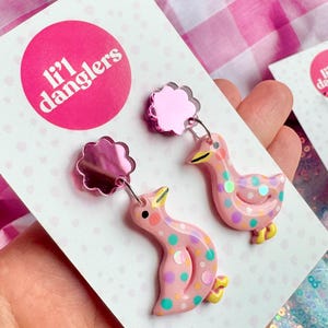 May include: A pair of pink duck earrings with colourful polka dots and yellow feet. The earrings are attached to a pink, cloud-shaped backing. The earrings are displayed on a white card with the text "li'l danglers" in a pink circle.