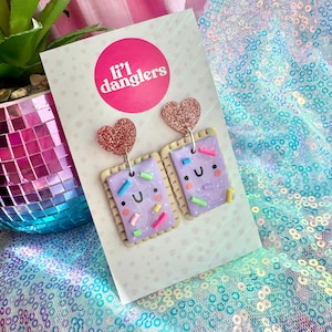 May include: A pair of Lil' Dangler's earrings. Each earring is a purple rectangle with a smiling face and colourful sprinkles, attached to a glittery pink heart stud. The background features a pink and white checked pattern and a sequined fabric.