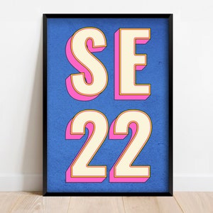 Peut inclure: Une impression encadrée avec le texte "SE 22" en grandes lettres blanches en 3D avec des contours roses et jaunes sur un fond bleu.