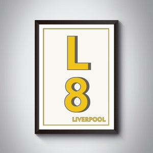 May include: Framed art print featuring the letter "L" above the number "8" in yellow, with a gray shadow effect. The word "LIVERPOOL" is printed in a smaller font below the number. The artwork is set against a cream background and framed in a dark brown frame.