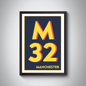 Op de afbeelding: Een ingelijst afdruk met een gele "M" en "32" op een donkerblauwe achtergrond. De tekst "MANCHESTER" staat onderaan de afdruk.