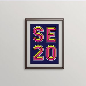 Puede incluir: Una impresión enmarcada con el texto "SE20" en una fuente geométrica en negrita. Las letras están delineadas en negro y rellenas de rosa y amarillo.