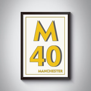 Op de afbeelding: Een ingelijst print met een gele "M" en "40" in een vet, sans-serif lettertype. De tekst "MANCHESTER" staat onder de nummers in een kleiner lettertype.