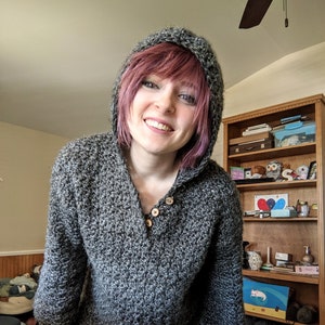 May include: A person wearing a gray hooded sweater with a button closure. The sweater has a speckled pattern and is made of a soft, cozy material.
