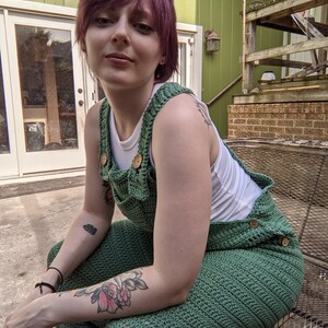 May include: A person wearing a green crocheted jumpsuit with wooden buttons. The person has short, purple hair and is sitting on a metal bench.