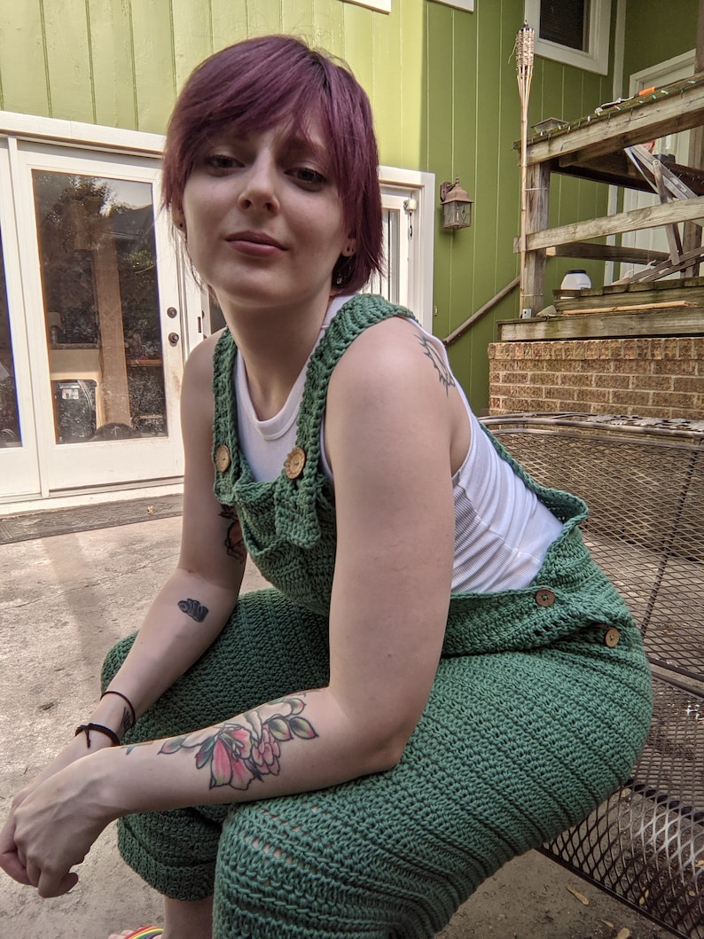 May include: A person wearing a green crocheted jumpsuit with wooden buttons. The person has short, purple hair and is sitting on a metal bench.