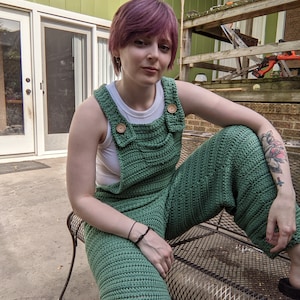 May include: A person wearing a green crocheted jumpsuit with a white tank top underneath. The person has short, pink hair and is sitting on a metal bench.
