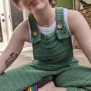 May include: A person wearing green crocheted overalls with rainbow striped slides. The person has a tattoo of an axolotl on their arm.