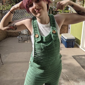 May include: A person wearing a green crocheted jumpsuit with brown buttons. The jumpsuit has a white tank top underneath. The person is flexing their biceps.