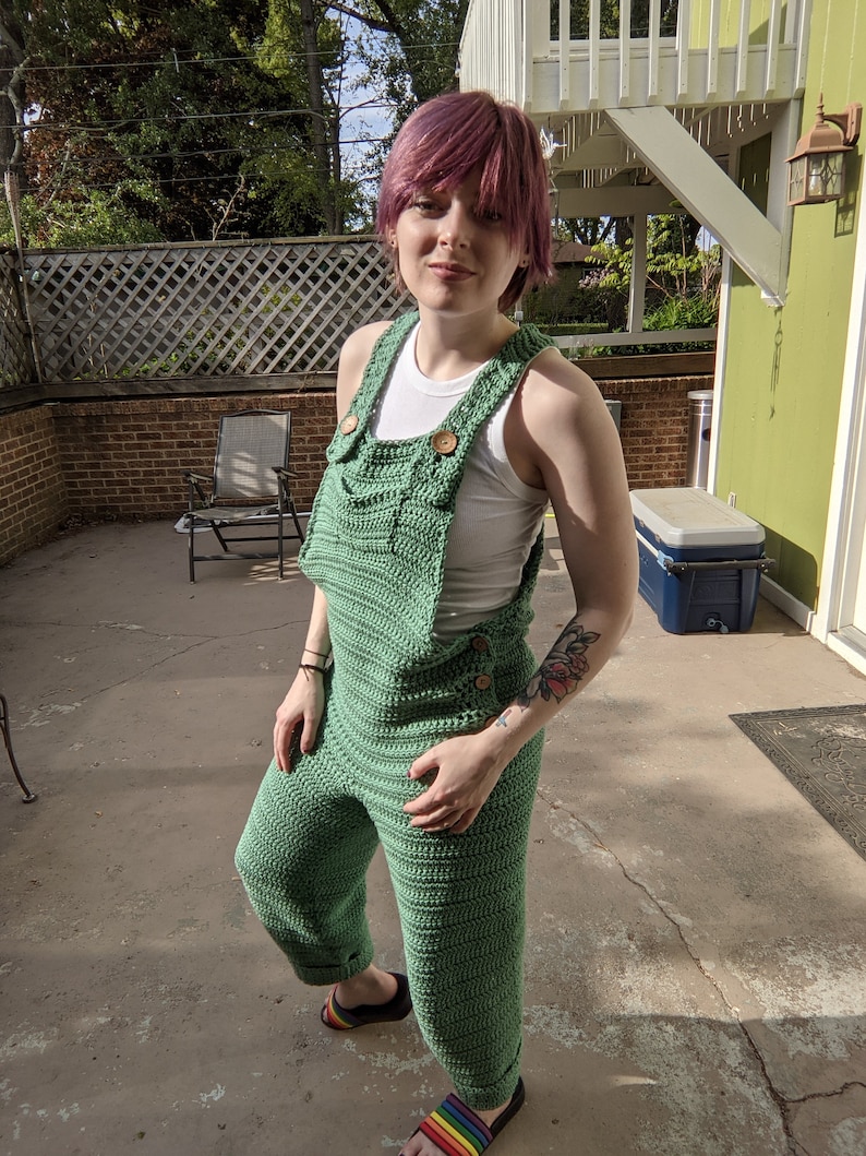 May include: A person wearing a green crocheted jumpsuit with brown buttons. The jumpsuit has a wide leg and a relaxed fit. The person is standing on a concrete patio.