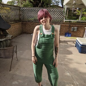 May include: A person wearing a green crocheted jumpsuit with rainbow sandals. The person has pink hair and is standing on a concrete patio.