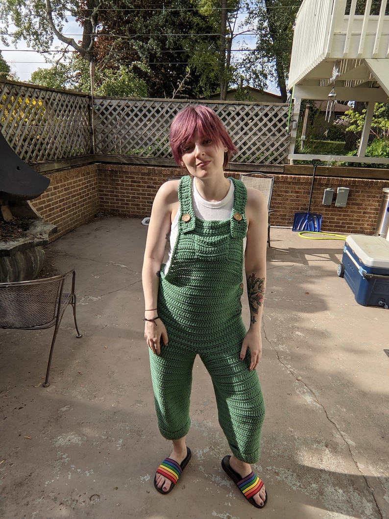May include: A person wearing a green crocheted jumpsuit with rainbow sandals. The person has pink hair and is standing on a concrete patio.