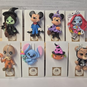May include: A collection of eight decorative character pins featuring various figures in Halloween-themed costumes, including a witch, a vampire, a skeleton, and a bear, each mounted on an individual white display card with a small sewing machine logo.