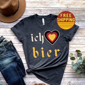 May include: A dark gray t-shirt with the text "ich &hearts; bier" in yellow and black lettering. The heart is red, yellow, and black. The shirt is on a wood background with a hat, jeans, and sandals.