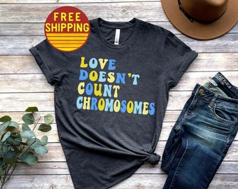 Puede incluir: Una camiseta gris oscuro con el texto "LOVE DOESN'T COUNT CHROMOSOMES" en letras de colores. La camiseta se combina con jeans azules, sandalias marrones y un sombrero marrón.