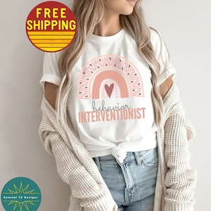May include: White t-shirt with a pink and peach rainbow design and a pink heart. The text "behavior interventionist" is printed below the rainbow.