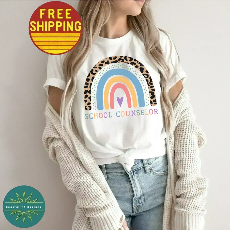 May include: A white t-shirt with a rainbow design and the text "SCHOOL COUNSELOR". The rainbow is made of colorful stripes with a leopard print border. A small purple heart is in the center of the rainbow.