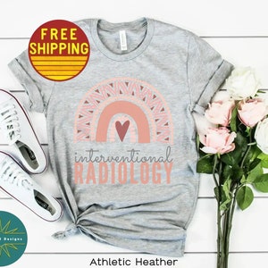 May include: Gray t-shirt with a pink and peach rainbow design and a heart. The text "interventional RADIOLOGY" is printed below the rainbow. The shirt is on a white surface with a pair of white sneakers and a bouquet of pink and white roses.