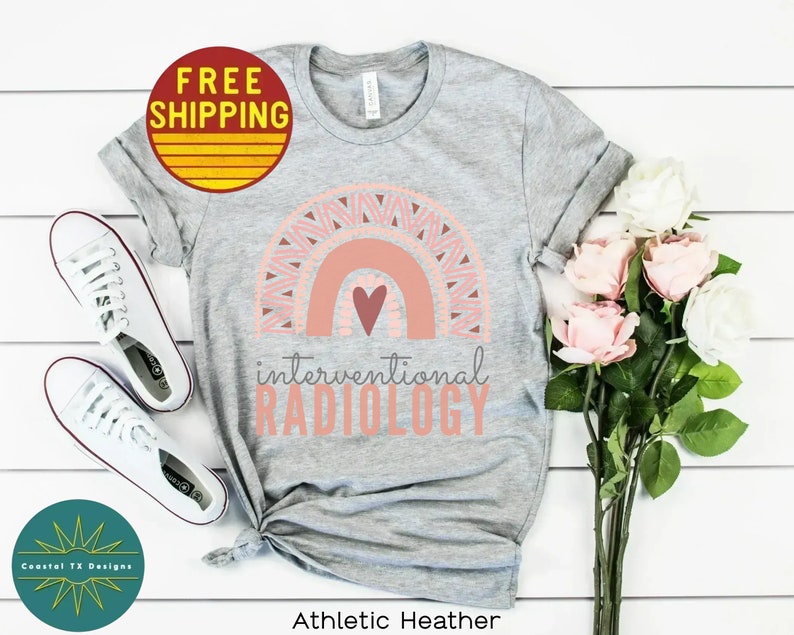 May include: Gray t-shirt with a pink and peach rainbow design and a heart. The text "interventional RADIOLOGY" is printed below the rainbow. The shirt is on a white surface with a pair of white sneakers and a bouquet of pink and white roses.
