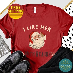 Op de afbeelding: Rood T-shirt met een vintage illustratie van de kerstman die een peperkoekhuis vasthoudt. De tekst "Ik hou van mannen met baarden" staat op het T-shirt gedrukt.