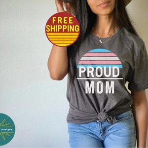 K&ouml;nnte beinhalten: Ein dunkelgraues T-Shirt mit einer wei&szlig;en Grafik eines Sonnenuntergangs mit einem Farbschema der Transgender-Pride-Flagge. Der Text "PROUD MOM" ist unterhalb der Sonnenuntergangsgrafik gedruckt.