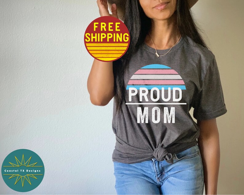 K&ouml;nnte beinhalten: Ein dunkelgraues T-Shirt mit einer wei&szlig;en Grafik eines Sonnenuntergangs mit einem Farbschema der Transgender-Pride-Flagge. Der Text "PROUD MOM" ist unterhalb der Sonnenuntergangsgrafik gedruckt.