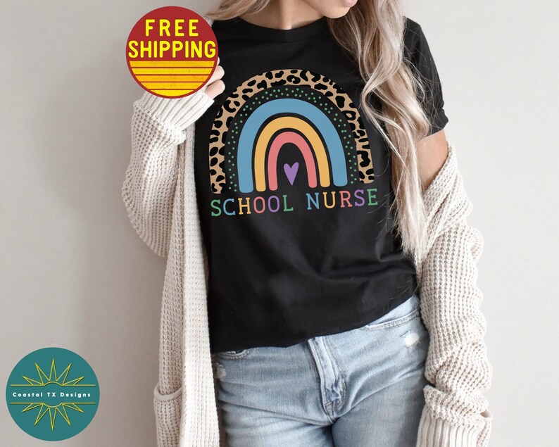Puede incluir: Una camiseta negra con un dise&ntilde;o de arco&iacute;ris e estampado de leopardo. El texto "SCHOOL NURSE" est&aacute; impreso debajo del arco&iacute;ris.