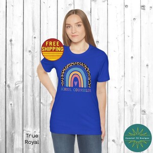 May include: A royal blue t-shirt with a rainbow and leopard print design. The text "SCHOOL COUNSELOR" is printed below the rainbow. The t-shirt is on a model.