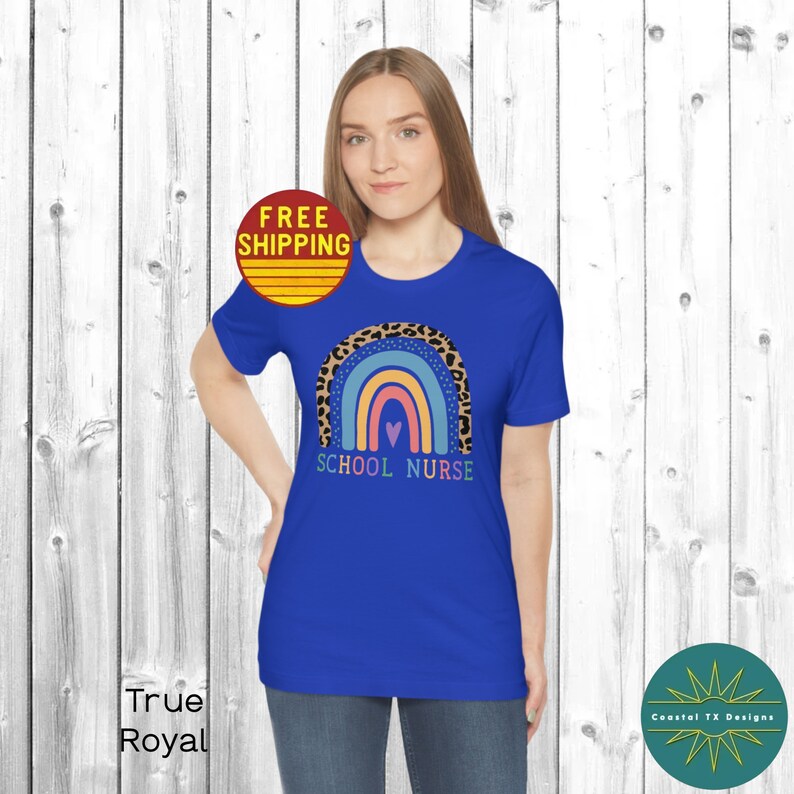 Puede incluir: Una camiseta azul real con un dise&ntilde;o de arco&iacute;ris con un borde de estampado de leopardo. El arco&iacute;ris tiene un coraz&oacute;n rosa en el centro y el texto "SCHOOL NURSE" debajo.